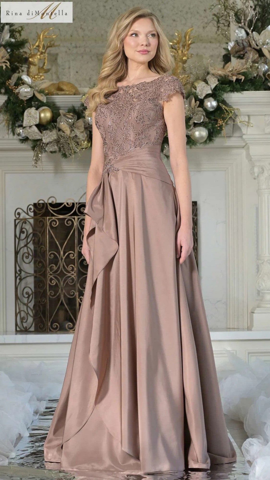 A woman in the mocha Rina Di Montella RD3327 formal gown, featuring a lace-embroidered bodice and floor-length skirt, stands before a festive fireplace decorated with greenery and ornaments.